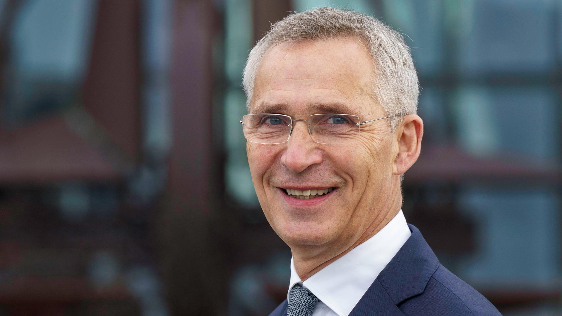 NATO-Generalsekretär Jens Stoltenberg steht vor einem Bürogebäude und lächelt in die Kamera.