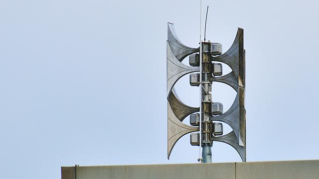 Auf einem Dach ist eine Sirenenanlage. Sie besteht aus mehreren Sirenen aus Metall, die in zwei Richtungen zeigen.