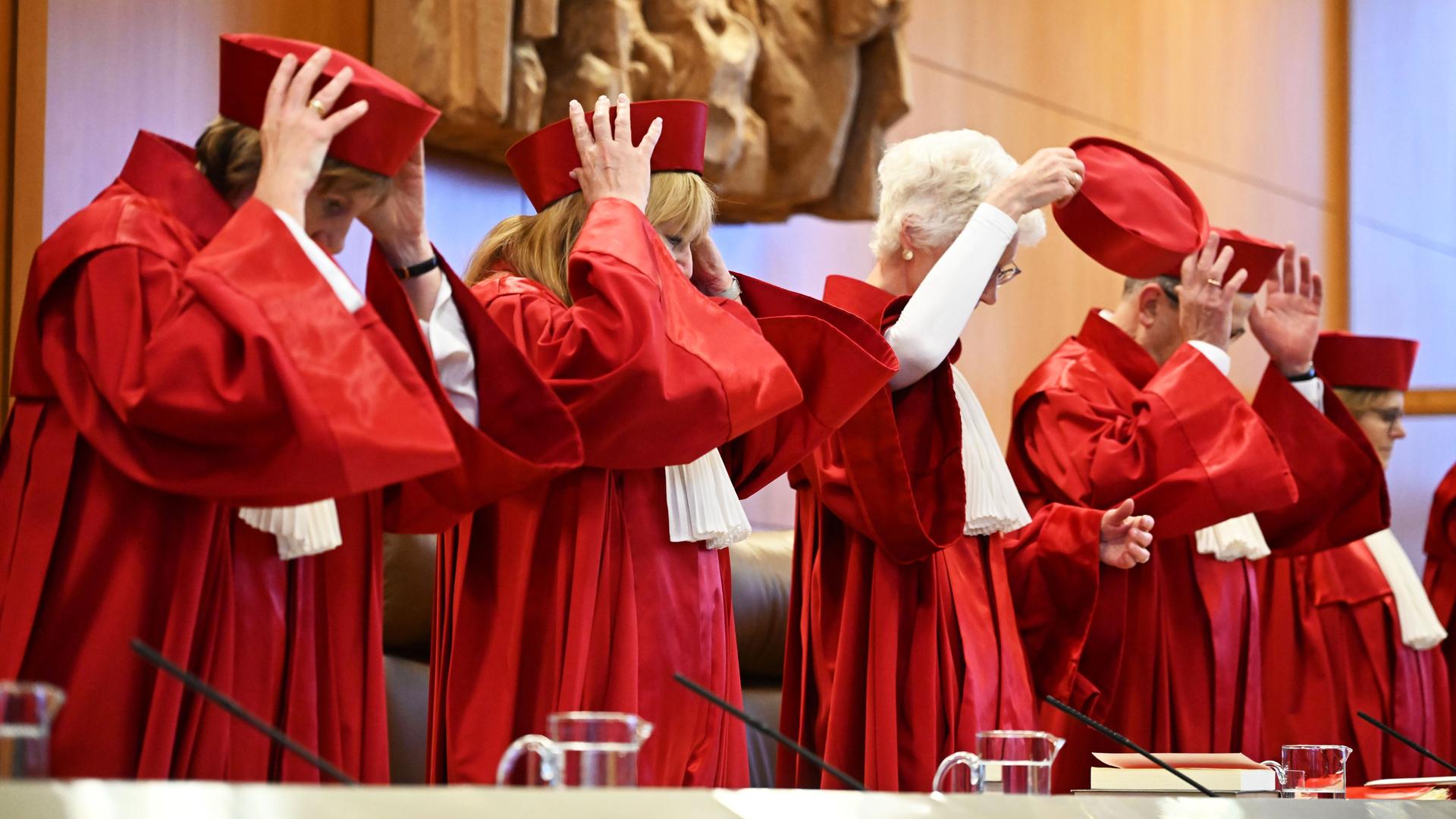 Richterinnen und Richter des Bundesverfassungsgerichts stehen vor einer Verhandlung in roten Roben hinter ihrem Tisch und setzen sich rote Mützen auf.