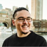 Headshot photograph of Nick DeJesus in front of a city skyline. He is smiling and has his hair slicked back into a ponytail.