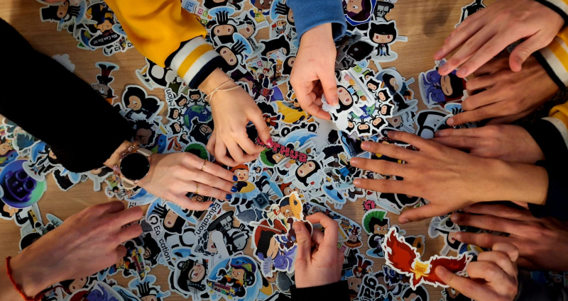 Several people's hands are reaching into a big pile of stickers on top of a table.