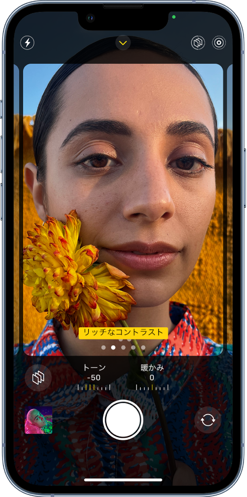 「カメラ」が開いていて、「フォトグラフスタイル」画面が表示されています。画面の左上隅には、フラッシュボタンがあります。画面の上部中央にはカメラコントロールボタンがあり、オンになっています。右上隅には、オンになっているフォトグラフ・スタイル・ボタンとLive Photosボタンがあります。画面いっぱいのカメラの枠に、「リッチなコントラスト」スタイルが表示されています。カメラの枠の下には、スタイルボタン、「トーン」と「暖かみ」の値を調整するスライダ、スタイル・リセット・ボタンがあります。画面の下部には、左から順に、写真およびビデオビューアボタン、写真撮影ボタン、背面カメラ選択ボタンがあります。