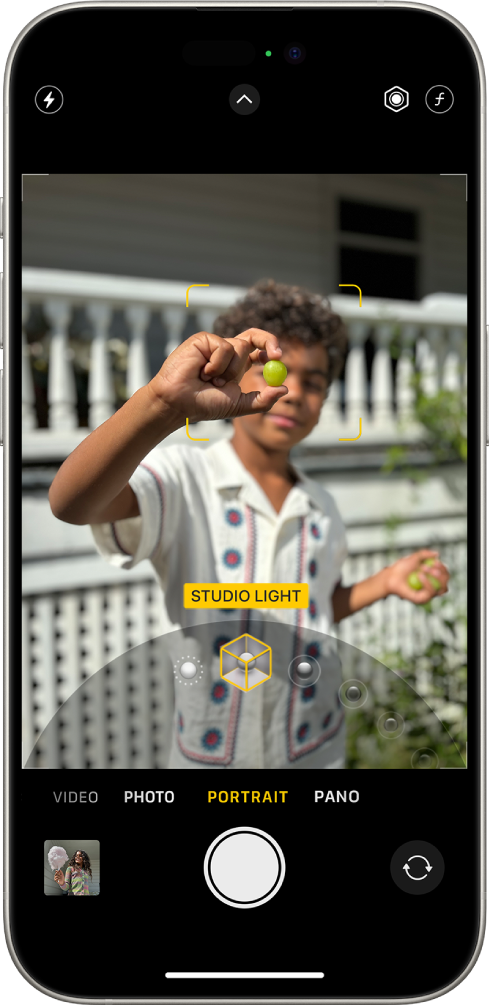 The Camera screen in Portrait mode; in the viewfinder, the subject is sharp and the background is blurred. The dial to select Portrait Lighting is open in the bottom of the frame and Studio Light is selected. At the top left of the screen is the Flash button, at the top center is the Camera Controls button, and at the top right are the buttons to adjust Portrait Lighting intensity and Depth Control. At the bottom of the screen are, from left to right, the Photo and Video Viewer button, the Take Picture button, and the Camera Chooser Back-Facing button.