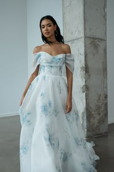 a woman in a white and blue dress standing next to a columned wall wearing an off the shoulder gown