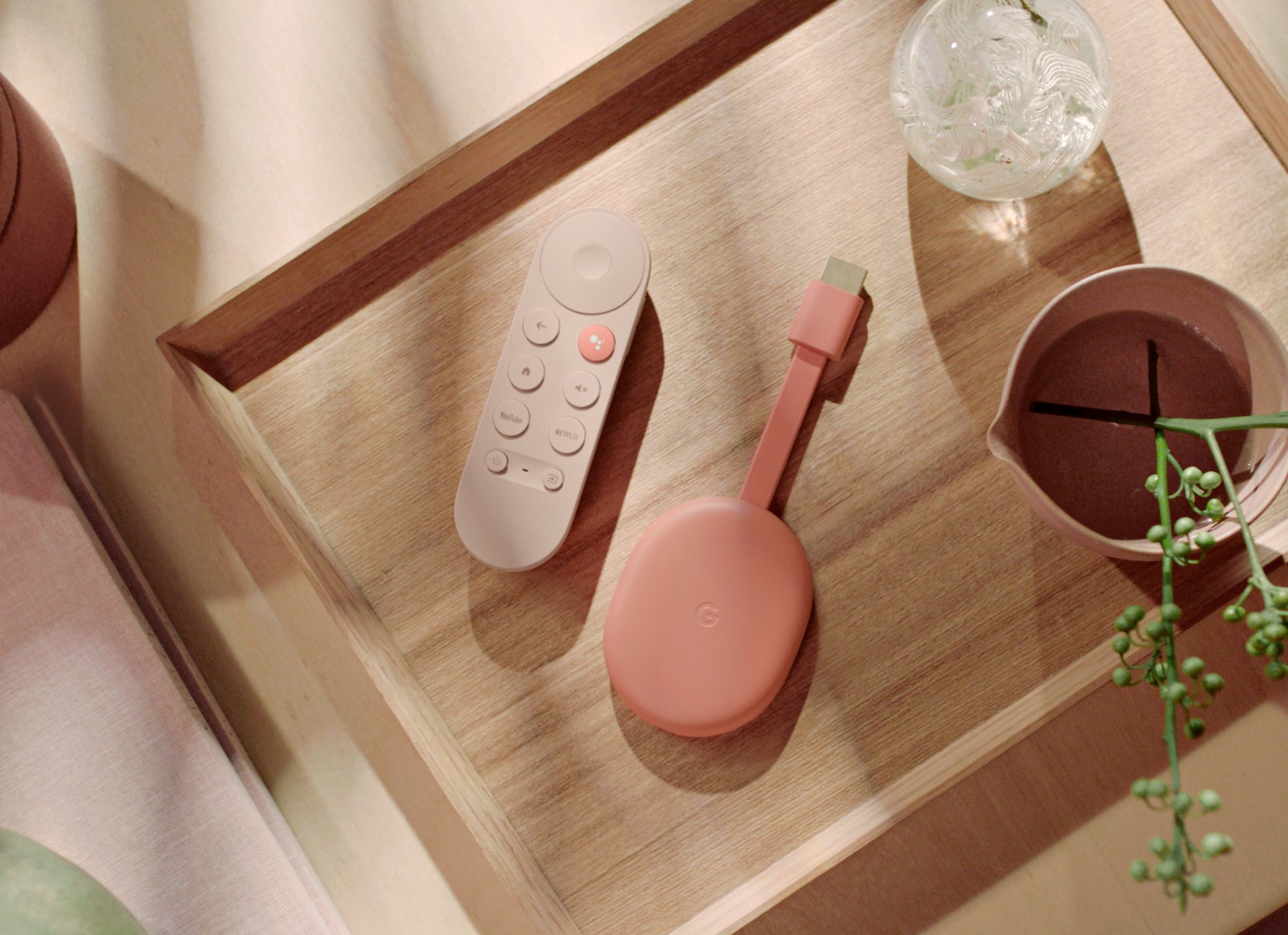 Chromecast with Google TV in Sunrise on a smooth wooden surface next to a plant and a glass art piece