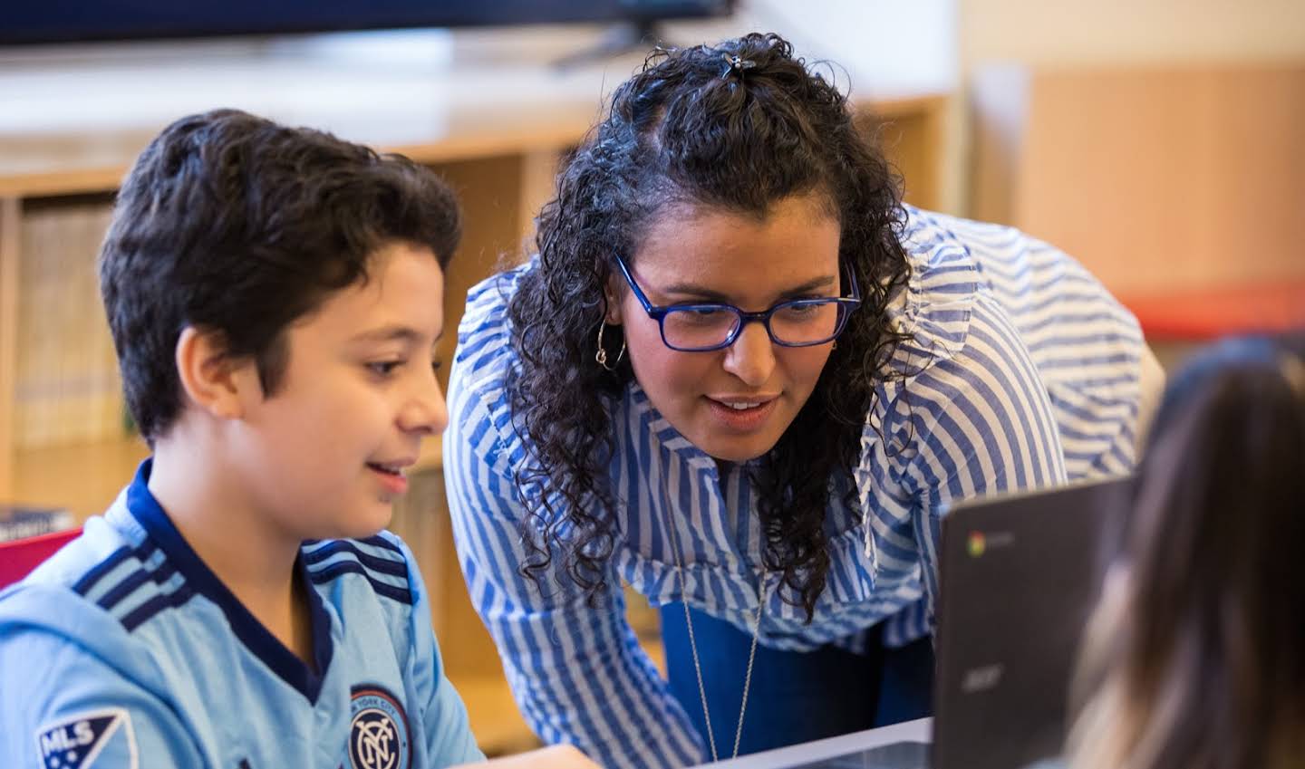 A close up of an educator with glasses leans over to review something on a student’s screen.