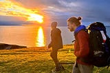 Two hikers walk against a fiery sunset over the sea.