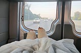 Two legs in thick casts under a blanket are seen from the inside of a hospital van. The highway stretches out beyond the back windows.