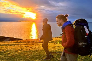 Two hikers walk against a fiery sunset over the sea.