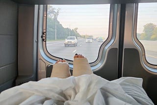 Two legs in thick casts under a blanket are seen from the inside of a hospital van. The highway stretches out beyond the back windows.