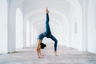 A woman dressed in blue leggings and tank top is doing a backbend amidst a gorgeous series of white archways. She has one leg high in the air.