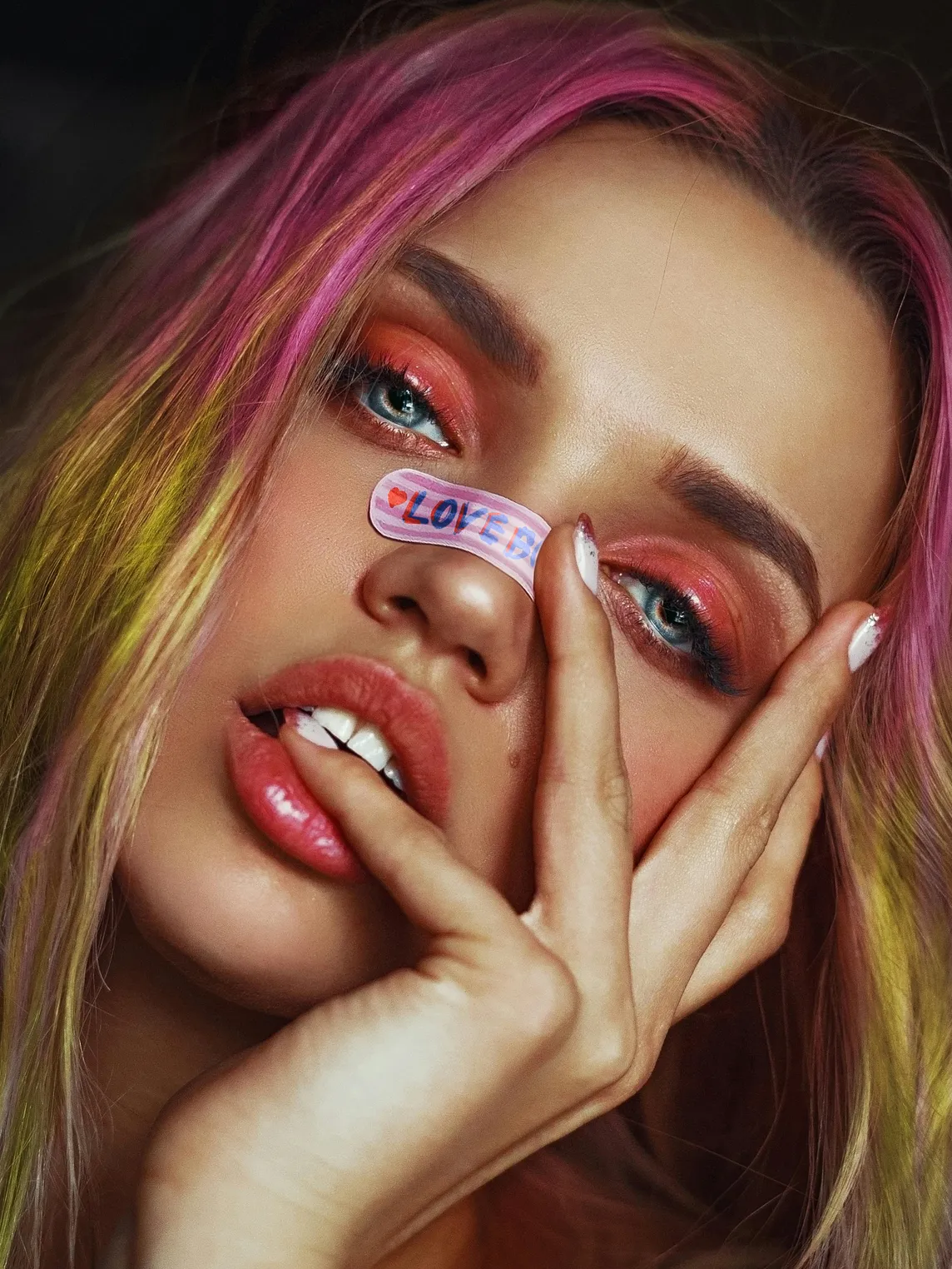 An airbrushed image of a woman with blue eyes and orange eyeshadow poses with her hand across her face. Her hair has pink roots and yellow ends, and a sticker over her nose reads “love”.