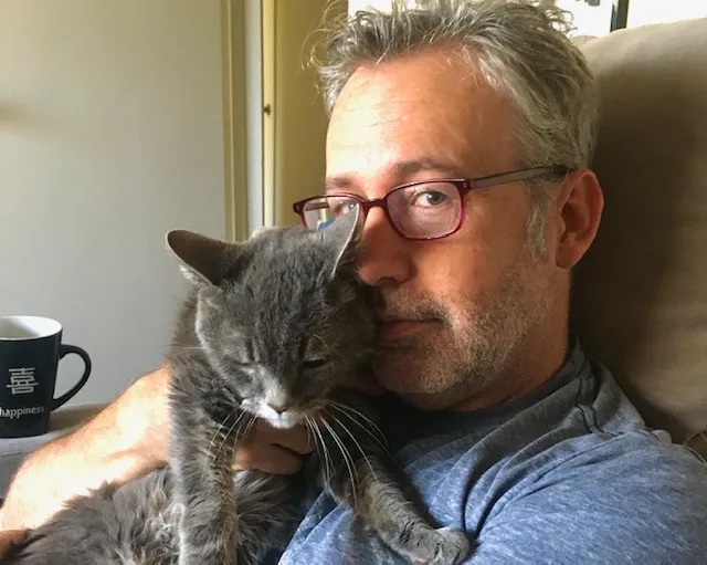 A man wearing red glasses snuggles with a gray cat.