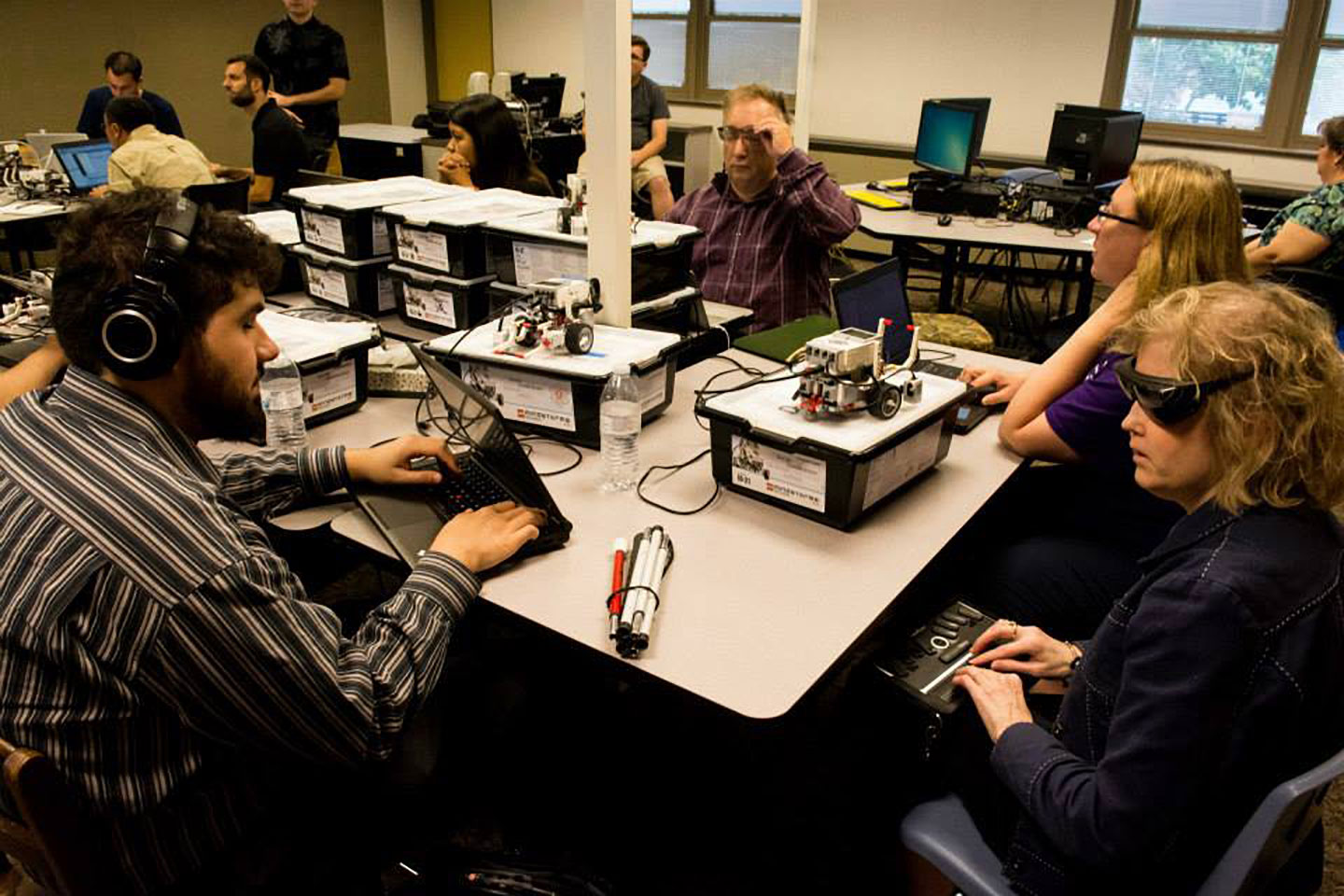 Sina sits at a table wearing headphones and hands on his laptop keyboard, next to many other people at computers and other devices. The tables are full of boxes and small robotic devices.