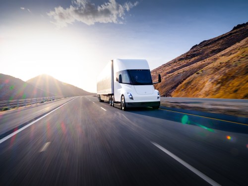 A white Tesla Semi truck is on a highway with a hill behind it.