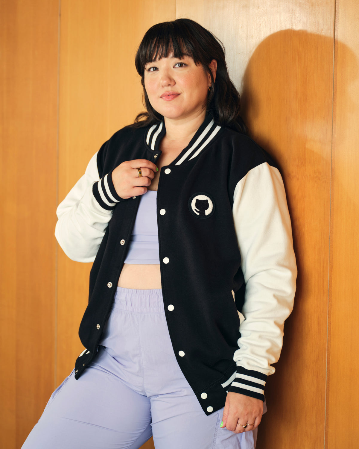 Photo of woman with black hair, posing wearing Mona Varsity Jacket over violet jumpsuit in office.