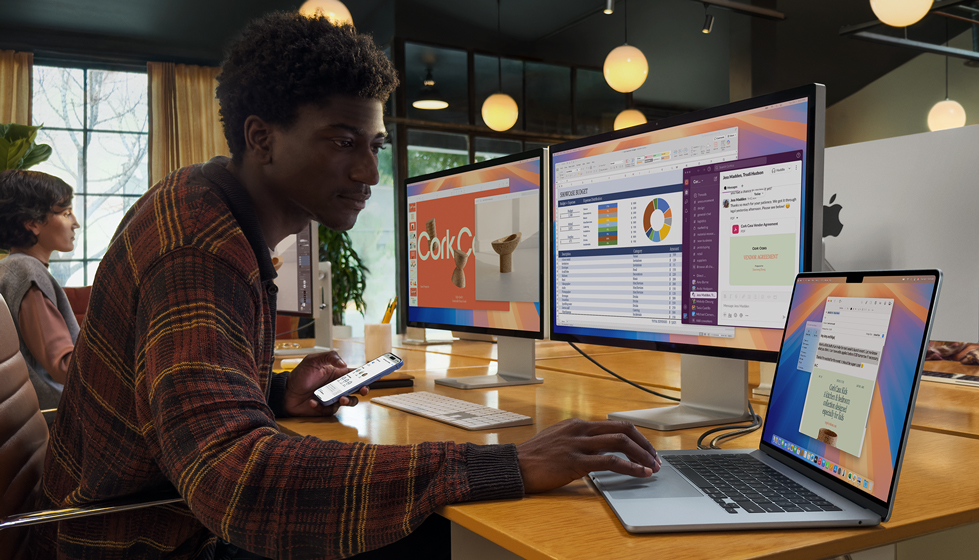 A person in an office environment using open MacBook Air with two external displays and Magic Keyboard, with iPhone in their other hand