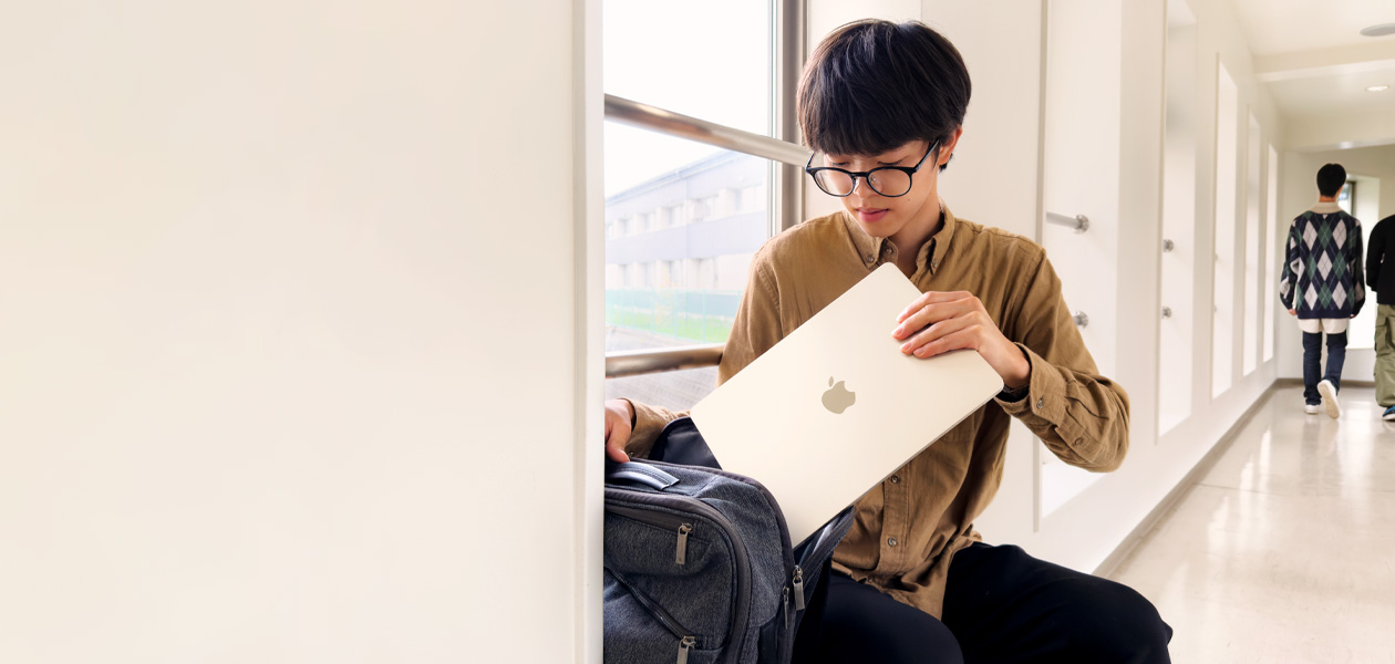 大学校内の廊下で窓際に座っている学生がバックパックからMacBook Airを取り出している。