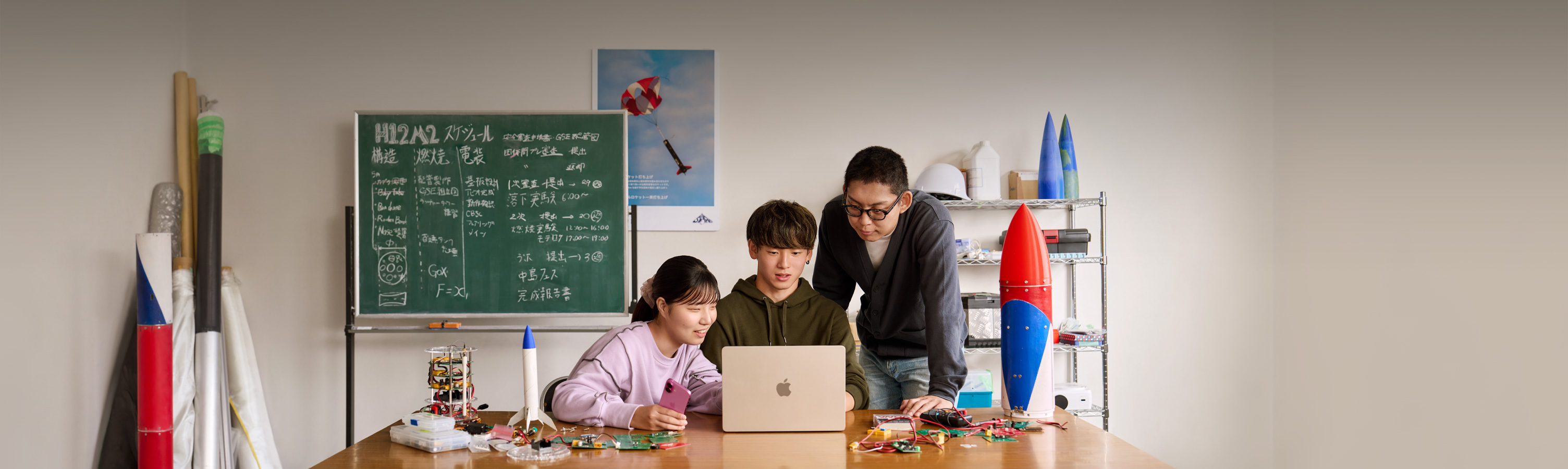 大学のロケットサークルの部室で3人の学生がMacBook Airを見ながら作業している。