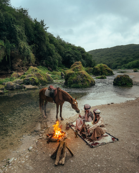 A Bedouin father and son sit on a rug next to a hot spring in Salalah, Oman, brewing coffee over a fire as their horse looks on.