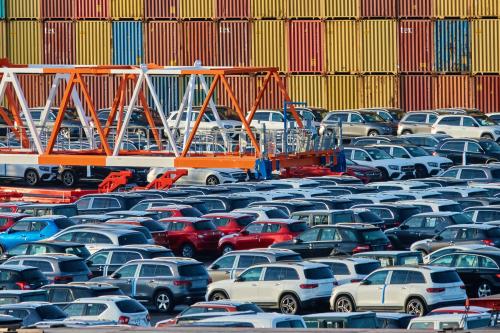 A parking lot full of new cars in the foreground. A stack of shipping containers in the background.