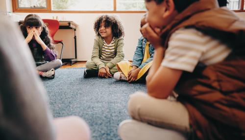 A group of children sit in a circle in a classroom listening to their teacher during a lesson.