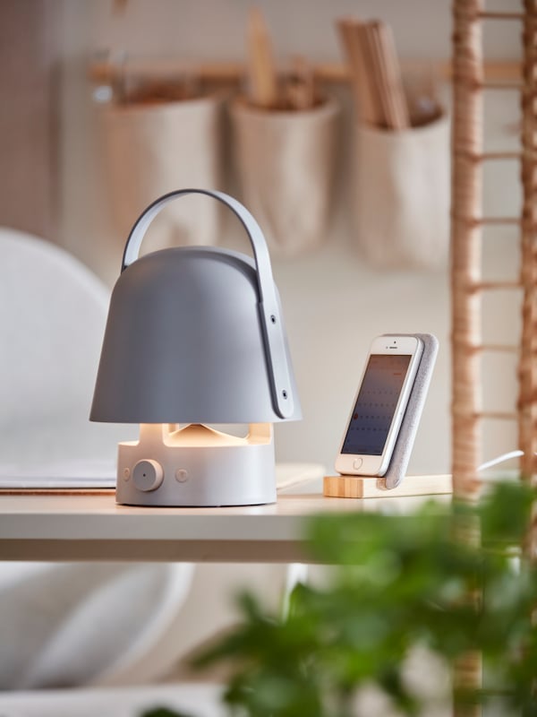 A grey VAPPEBY Bluetooth speaker lamp is placed on a white desk, next to a NORDMÄRKE wireless charging stand made of bamboo.