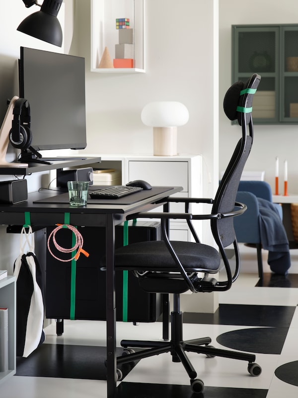 A side view of a PC gaming setup with a black UTVISNING gaming desk with shelf and a dark grey chair in a white passageway.