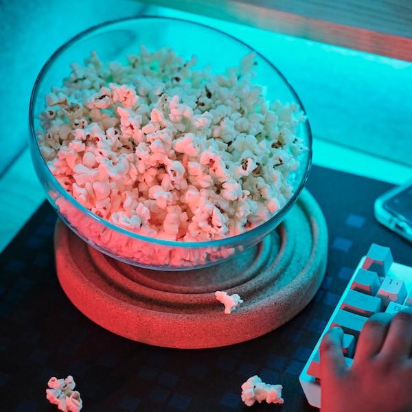 A BRÄNNBOLL coaster is set under a glass bowl full of popcorn on a desk. A BRÄNNBOLL gaming mouse pad is under the coaster.