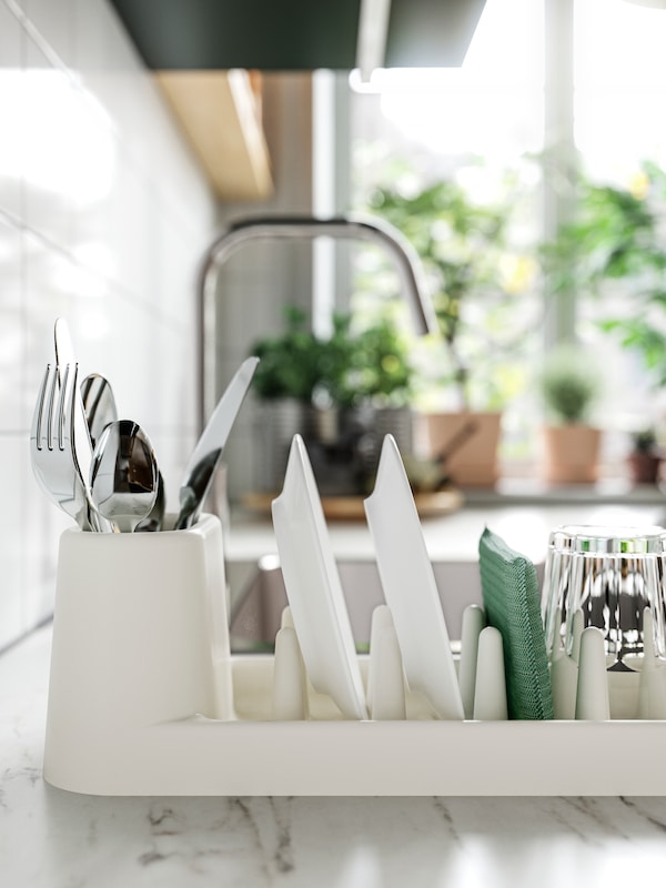 An off-white STÄMLING dish drainer holding a glass, two white plates and cutlery, on a white marble effect/laminate worktop.