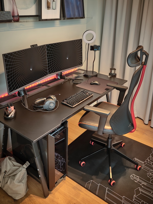 A black MATCHSPEL gaming chair and UTESPELARE gaming desk with a grey curtain on one side and two monitors on top.