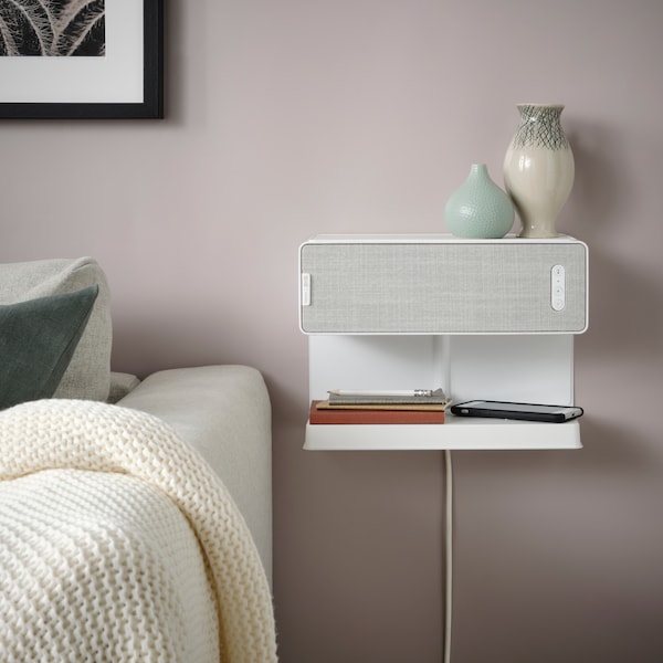 A white SYMFONISK Wi-Fi bookshelf speaker is hanging on the wall in a living room next to a white sofa.