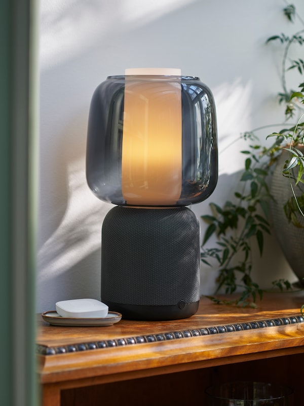 A black SYMFONISK speaker lamp base with Wi-Fi and a black glass shade is placed on a wooden table by a green plant.