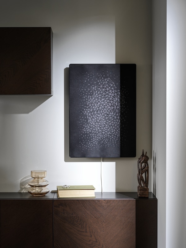 A black SYMFONISK picture frame with Wi-Fi speaker is hanging on a white wall over a dark brown cabinet.