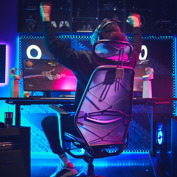 A dark gaming room with a lit-up gaming station. A person, sitting in a gaming chair, is raising their arms in victory.