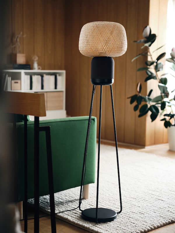 A SYMFONISK floor lamp with Wi-Fi speaker is standing in a living room next to a green sofa on a cream rug.