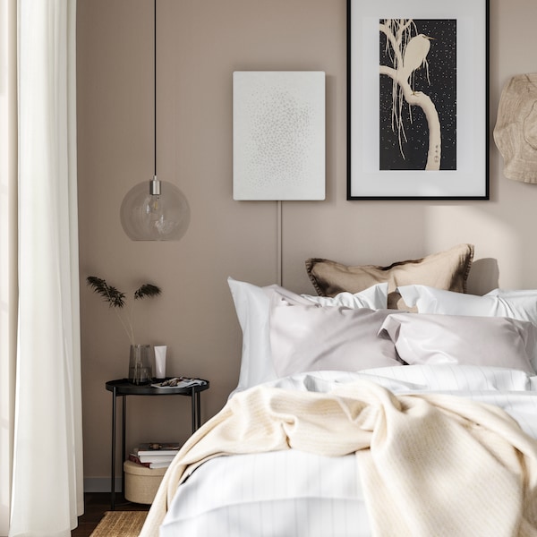 A white SYMFONISK picture frame with Wi-Fi speaker hangs on a beige bedroom wall behind a bed with white bedlinen.