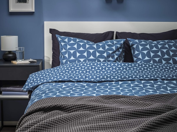 A bed made with a geometric pattern duvet cover in blue and a dark gray blanket at the bottom of the bed.