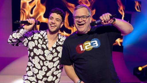 "1, 2 oder 3 - Popstars on Tour": Luca Hänni singt ins Mikrofon. Elton steht dabei. Hinter ihnen sind Flammen zu sehen.