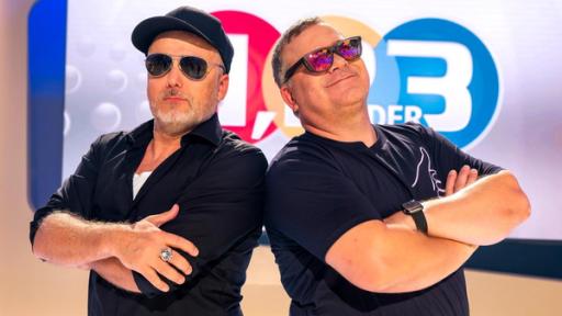 1, 2 oder 3: Moderator Elton und Studiogast stehen vor der Vidiwall im "1, 2 oder 3"-Studio mit verschränkten Armen und Sonnenbrillen.